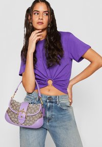 Lilla cropped t-shirt med en tvistet frontdetalje, parret med en lavendel- og beige håndtaske, der har en guldbuckle og mønsterdesign.