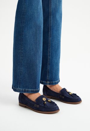Mocassini in suede blu navy con nappine, suola piatta e dettagli decorativi in oro. Indossati con pantaloni larghi in denim blu.