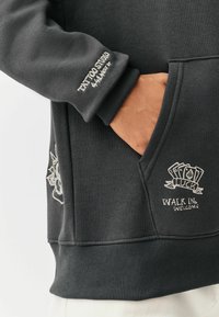 Donkergrijze sweatshirt met ribgebreide mouwen en zoom, met witte geborduurde opdrukken: "Tattoo Studio," "Luck," en "Walk Ins Welcome."