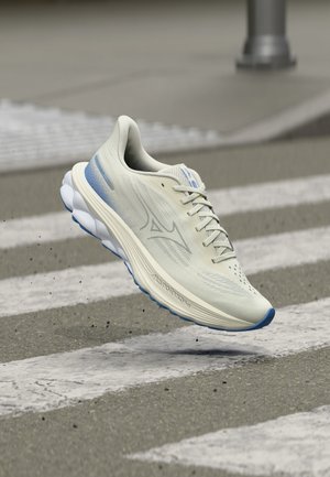 Chaussure de course blanche et bleue en l'air au-dessus d'un passage piéton sur une route asphaltée, montrant les détails du côté et de la semelle inférieure.