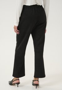 Pantalones de ante negro con corte relajado, que cuentan con una cinturilla, perneras acampanadas y una textura suave. Combinados con tacones de slingback negros.