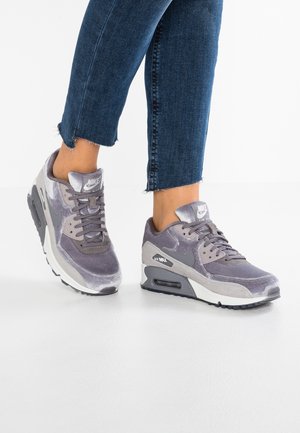 Person med grå Nike Air Max sneakers med hvide såler og mørkeblå afkortede jeans på et hvidt gulv.