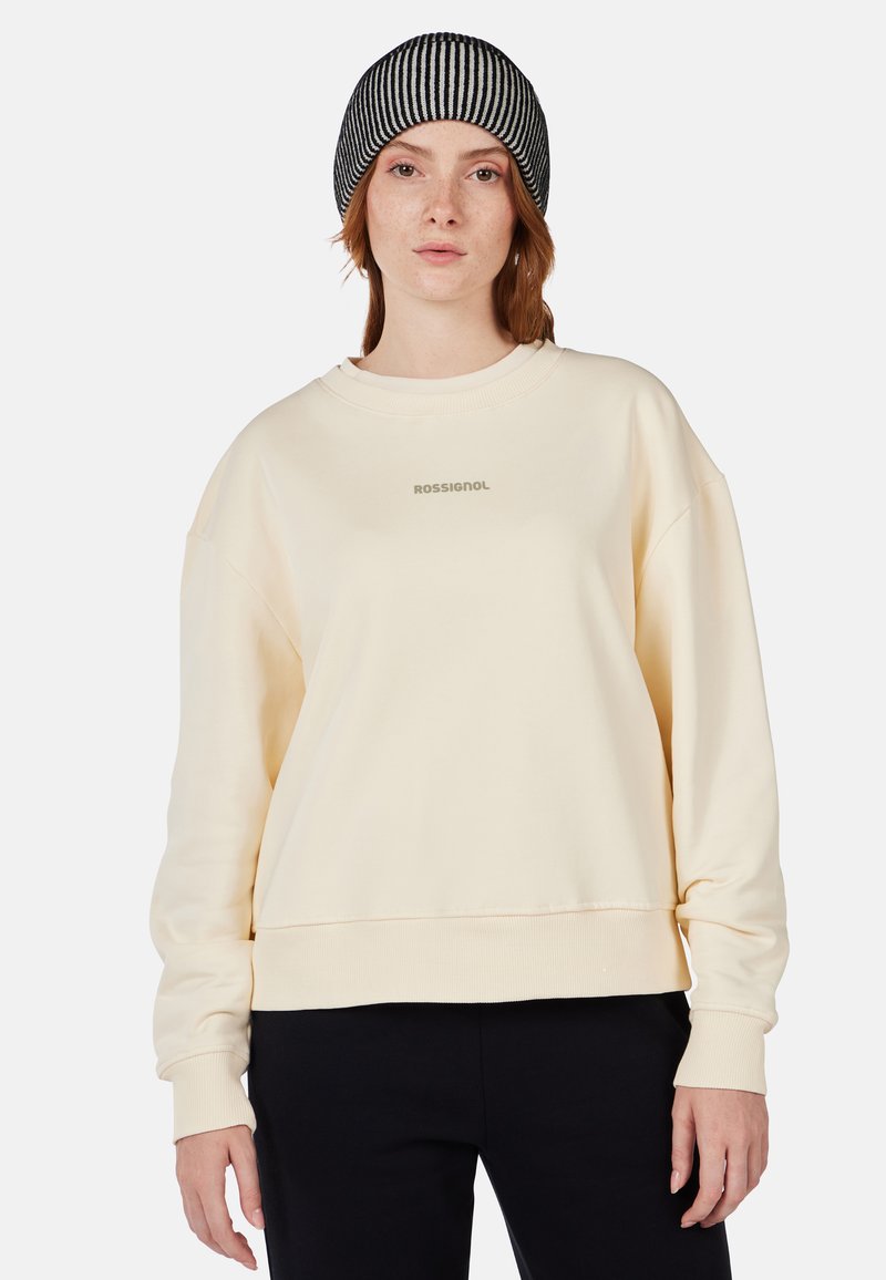 Sudadera color crema con hombros caídos, puños y dobladillo acanalados; presenta un pequeño logo centrado. Gorro de rayas blanco y negro en la cabeza.