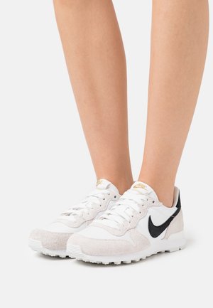 Valkoiset ja mustat Nike-urheilukengät, joissa on käytetty sekä mokkaa että verkkomateriaaleja, pyöreä kärki, nauhakiinnitys ja kultainen logo-aksentti kielen kohdalla.
