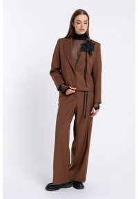 Brązowy garnitur skrojony na miarę z dopasowaną marynarką, przezroczystą czarną bluzką pod spodem oraz ozdobnym czarnym kwiatowym akcentem na klapie. Wśród detali znajdują się długie wstążki.