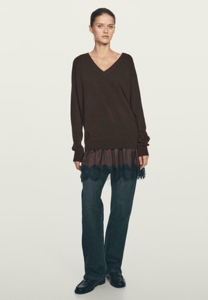 Donna che indossa un ampio maglione marrone scuro a V sopra una camicia con pizzo, jeans scuri e mocassini neri, in piedi su uno sfondo chiaro.