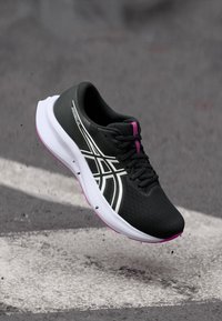 Μαύρο αθλητικό παπούτσι με υφασμάτινη επιφάνεια, λευκές και ροζ λεπτομέρειες, μαλακή σόλα και αεροδυναμικό σχέδιο, suspend πάνω από μια επιφάνεια.