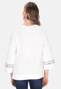 Vit långärmad topp med rund halsringning och utsvängda ärmar, med ett blommönstrat band som dekoration vid ärmsluten. Mjuk textur, avslappnad design.