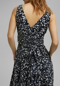 Kvinna i ärmläns klänning i marinblått med vit blomstertryck, djup V-back och knytdetalj i midjan, sedd bakifrån.