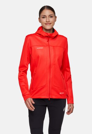 Kvinde iført en knaldrød Mammut hættejakke med lynlåslommer foran og på brystet, kombineret med sorte bukser, smilende mod hvid baggrund.