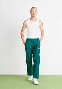 Groene sportbroek met witte strepen en Adidas-logo, gedragen met een witte tanktop en witte sneakers, op een lichte achtergrond.