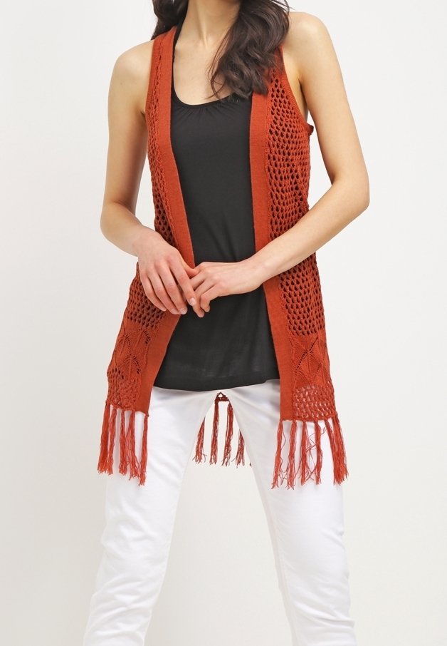 Rostfarbene gehäkelte Weste mit offenem Vorderteil, strukturierten Strickmustern und Fransen am Saum, kombiniert mit einem schwarzen Oberteil und weißen Hosen.