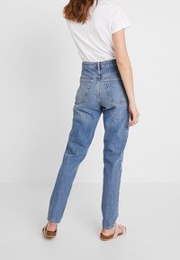 Korkeavyötäröiset siniset farkut, joissa on rento istuvuus, takataskut ja hienovarainen haalistunut pinta, yhdistettynä vaaleanruskeisiin sandaaleihin.