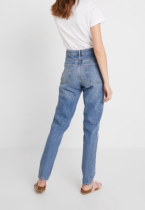 Jeans de tiro alto en denim azul con un corte relajado, que cuentan con bolsillos traseros y una sutil textura desgastada, combinados con sandalias de color tostado.