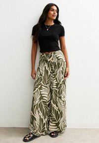 Black fitted t-shirt paired with loose, olive green trousers featuring large, cream foliage patterns. Sandals with a thick sole and accents.