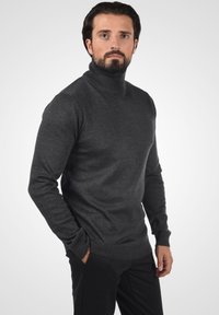 Suéter de cuello alto gris oscuro hecho de un material suave, con mangas largas y puños acanalados, combinado con pantalones negros con textura.