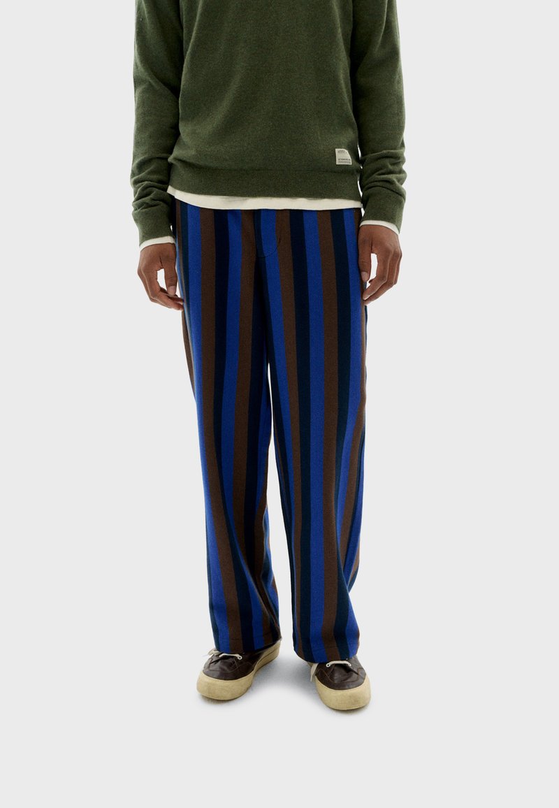 Pantalones de pierna ancha a rayas en azul, marrón y negro. El material parece suave; se combinan con un suéter verde y zapatos casuales. Diseño simple.