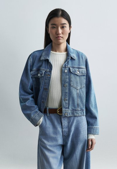 Jeune femme aux cheveux longs et lisses portant une veste en denim clair, un jean taille haute et un t-shirt blanc côtelé sur un fond uni.