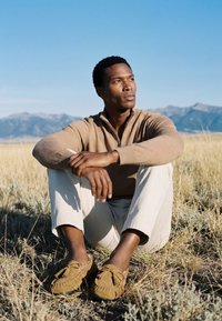 Jeune homme en pull beige et pantalon blanc assis dans de l'herbe sèche avec des montagnes et un ciel bleu clair en arrière-plan, regardant pensivement sur le côté.