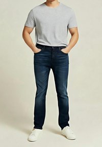 Camiseta gris de cuello redondo combinada con vaqueros azul oscuro. El modelo lleva zapatillas blancas. Los vaqueros tienen un ajuste slim y un desgaste sutil.