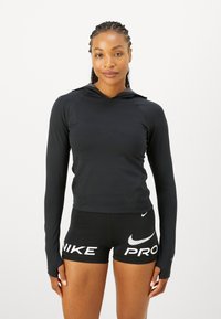 Melnas garās piedurknes džemperis ar pieguļošu dizainu, ar kapuci un bezšuvēm plecu apvidū. Apvienots ar melniem šortiem, uz kuriem redzams baltais "Nike Pro" logo.