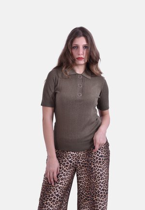 Donna con capelli mossi sciolti indossa una polo a maniche corte in maglia verde oliva e pantaloni ampi con stampa leopardata contro uno sfondo semplice.