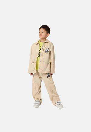 Beige Baumwolljacke und passende Hose mit Taschen; darunter grünes Hemd; mit schwarzen Stickereien und legerer Passform.