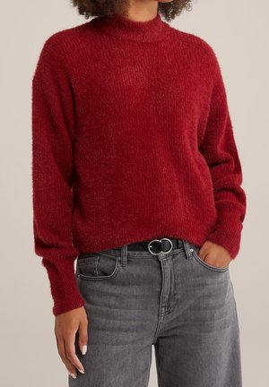Woman wearing a fuzzy red sweater with a high neckline tucked into high-waisted gray jeans with a black double-ring belt.