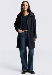 Marineblauer Mantel mit großem Kragen, Knöpfen vorne, strukturierter Stoff, getragen über einem dunklen Oberteil und blauen Jeans, mit einem hellbraunen Gürtel und schwarzen Schuhen.