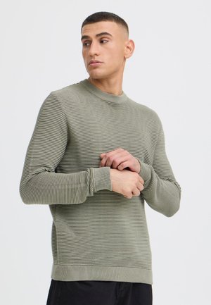 Jeune homme aux cheveux courts portant un pull côtelé vert olive clair, regardant vers sa gauche, mains jointes près de sa poitrine sur un fond uni.