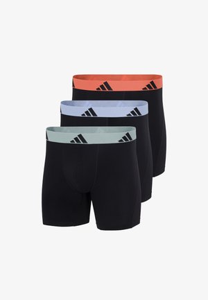 Atletyczny mężczyzna stojący boso, ubrany w czarne bokserki z pomarańczowym pasem w talii z czarnymi logo Adidas, na jednolitym tle.