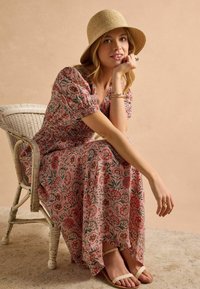 Florales Maxikleid mit Puffärmeln, rosa Grundfarbe, aufwendige rote und grüne Muster. Geflochtener Strohhut, gestapelte Armbänder und beigefarbene Sandalen.