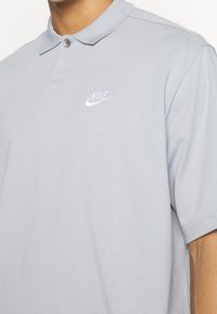 Light gray polo shirt made of cotton. Features a collared neck, two buttons, and a small white Nike logo on the left chest. Short sleeves.