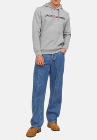 Grå sweatshirt med logo, forlommer og snøre-hætte, kombineret med løse blå jeans og brune sko.