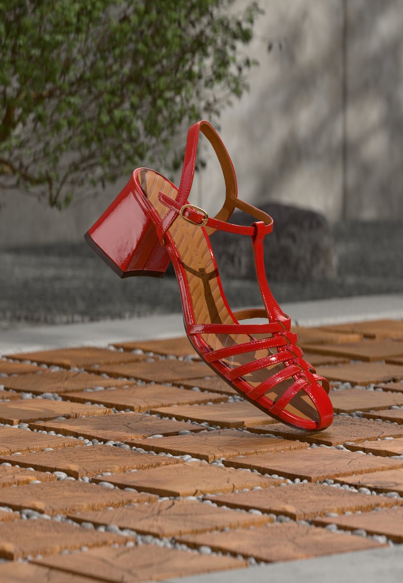 Κόκκινο σανδάλι από δερμάτινο βερνίκι με χοντρό τακούνι και λουράκι αστραγάλου που αιωρείται πάνω από ξύλινο πλακίδιο σε εξωτερικό χώρο.