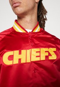 Giacca bomber in raso rosso con logo giallo "CHIEFS", colletto a coste gialle e bianche a strisce, e chiusura a bottoni a pressione.