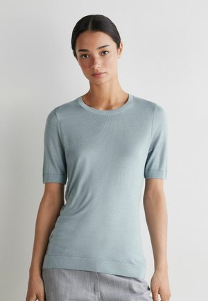 Jonge vrouw met donker haar, gekleed in een lichtblauw gebreid shirt met korte mouwen en grijze broek, staande tegen een witte muur.