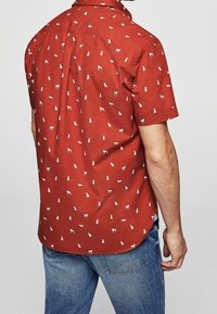 Homme portant une chemise à manches courtes couleur rouille avec des motifs de petits chiens blancs, associée à un jean bleu en denim, debout devant un fond uni.