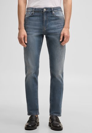 Homme portant un jean en denim bleu slim et des mocassins en cuir noir, debout devant un fond clair uni.