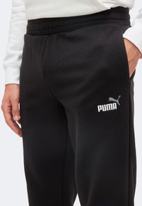 Άτομο που φορά μαύρο παντελόνι φόρμας Puma με λευκό λογότυπο στον αριστερό μηρό και λευκό φούτερ, με το αριστερό χέρι να ακουμπά πλάι του.