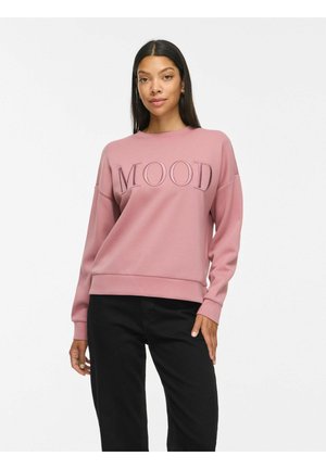 Femme aux longs cheveux foncés portant un sweat-shirt rose avec le mot "Mood" brodé sur le devant et un pantalon noir sur fond blanc.