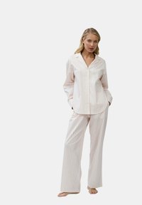 Frau steht barfuß in weißen Schlafanzug mit dünnen roten vertikalen Streifen und langen Ärmeln, Hände in den Taschen, vor einfarbigen Hintergrund.