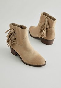 Stivaletti alla caviglia in suede beige con punta affilata, chiusura con zip laterale e dettagli in frange sul retro, poggiati su un tacco in legno.
