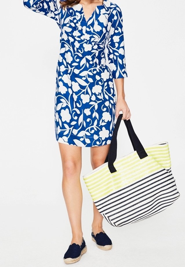 Blå och vit blommig klänning med trekvartsärmar; håller en stor randig strandväska med gula och svarta mönster. Espadrillos.