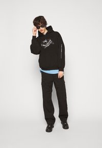 Schwarzer Hoodie mit grafischem Design, über einem hellblauen Shirt getragen. Schwarze Cargo-Hose, schwarze Schuhe und Sonnenbrille. Schlichter, urbaner Stil.