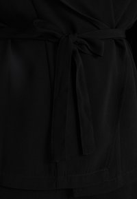 Černý obalový top z látky s uvázaným páskem v pase, hladkou texturou a minimalistickým designem s prošívaným lemem.