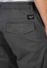 Pantalones cortos cargo grises con cintura elástica, bolsillo trasero y detalles de costura. Presenta una etiqueta negra con texto blanco en la cinturilla.