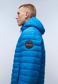 Blå puffjacka med huva, quiltat mönster och en framträdande logotyptryck på vänster ärm. Tyget verkar slätt och glänsande.