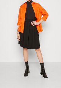 Oranžová košile na knoflíky, černý přiléhavý rolák, černá skladaná sukně a černé kotníkové boty s masivními podpatky. Prsten jako doplněk na ruce.