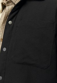 Zwarte stoffen shirt met een opvallende borstzak, drukknoopjes en een gestructureerd oppervlak. Gelaagd over een tan geruit shirt.